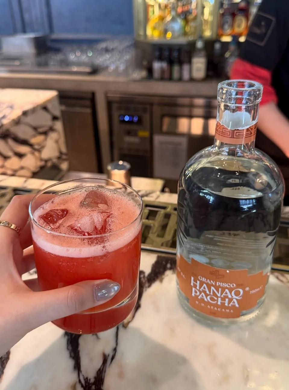 Coctel rojo junto a botella Hanaq Pacha en barra